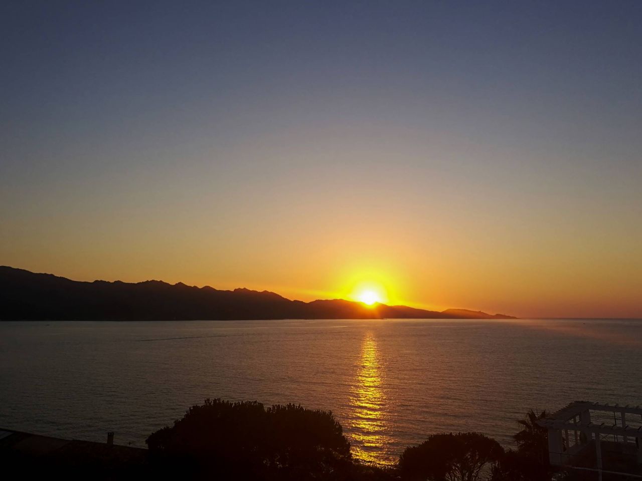 Un coucher de soleil serein sur des eaux calmes, avec des rayons dorés se reflétant sur la mer et des montagnes en silhouette en arrière-plan.