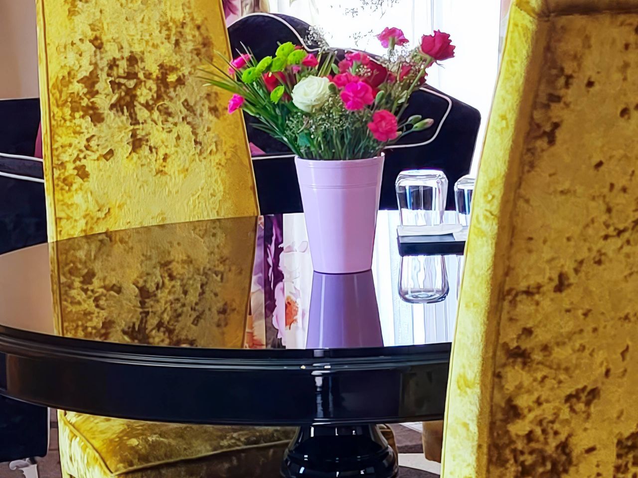 Une configuration de salle à manger moderne avec des chaises en velours jaune, une table noire et un arrangement floral coloré dans un vase rose.